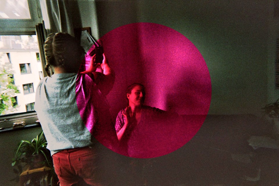 Bandet Rosa Huset foto med två personer, den ena personen har ryggen emot betraktaren och fotograferar den andra personen som sitter ner, i mitten av bilden är det en cerice cirkel som ligger i ett lager ovanför fotografiet av de två personerna.