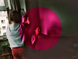Bandet Rosa Huset foto med två personer, den ena personen har ryggen emot betraktaren och fotograferar den andra personen som sitter ner, i mitten av bilden är det en cerice cirkel som ligger i ett lager ovanför fotografiet av de två personerna.