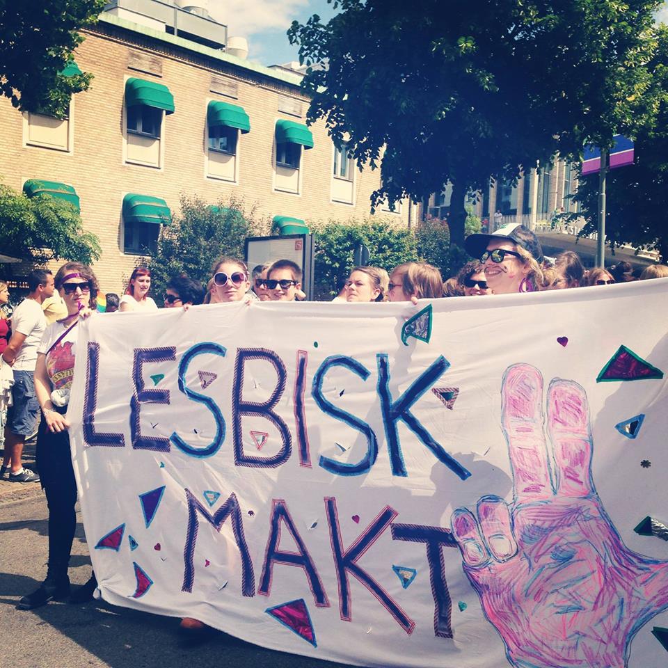 Några personer står bakom en banderoll i en parad. Banderollen är vi och på den står texten "Lesbisk Makt". Bredvid texten är en hand i en gest där mellan- och pekfinger samt tumme sträcks ut och de andra två fingrarna är vikta.